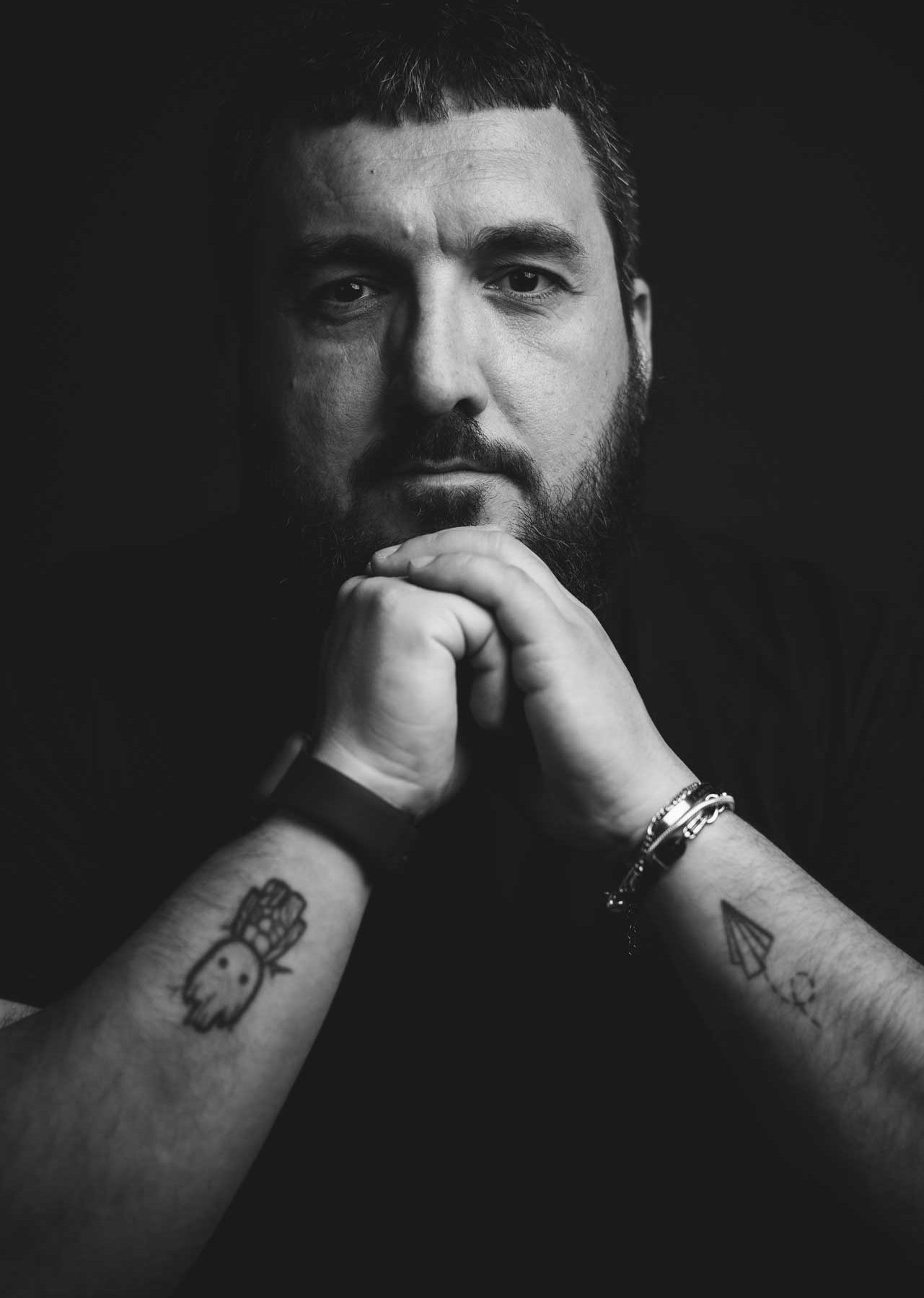Max Giubilei fotografo professionista – ritratto emotivo in bianco e nero Primo piano in bianco e nero di Max Giubilei, fotografo, con le mani giunte e tatuaggi visibili